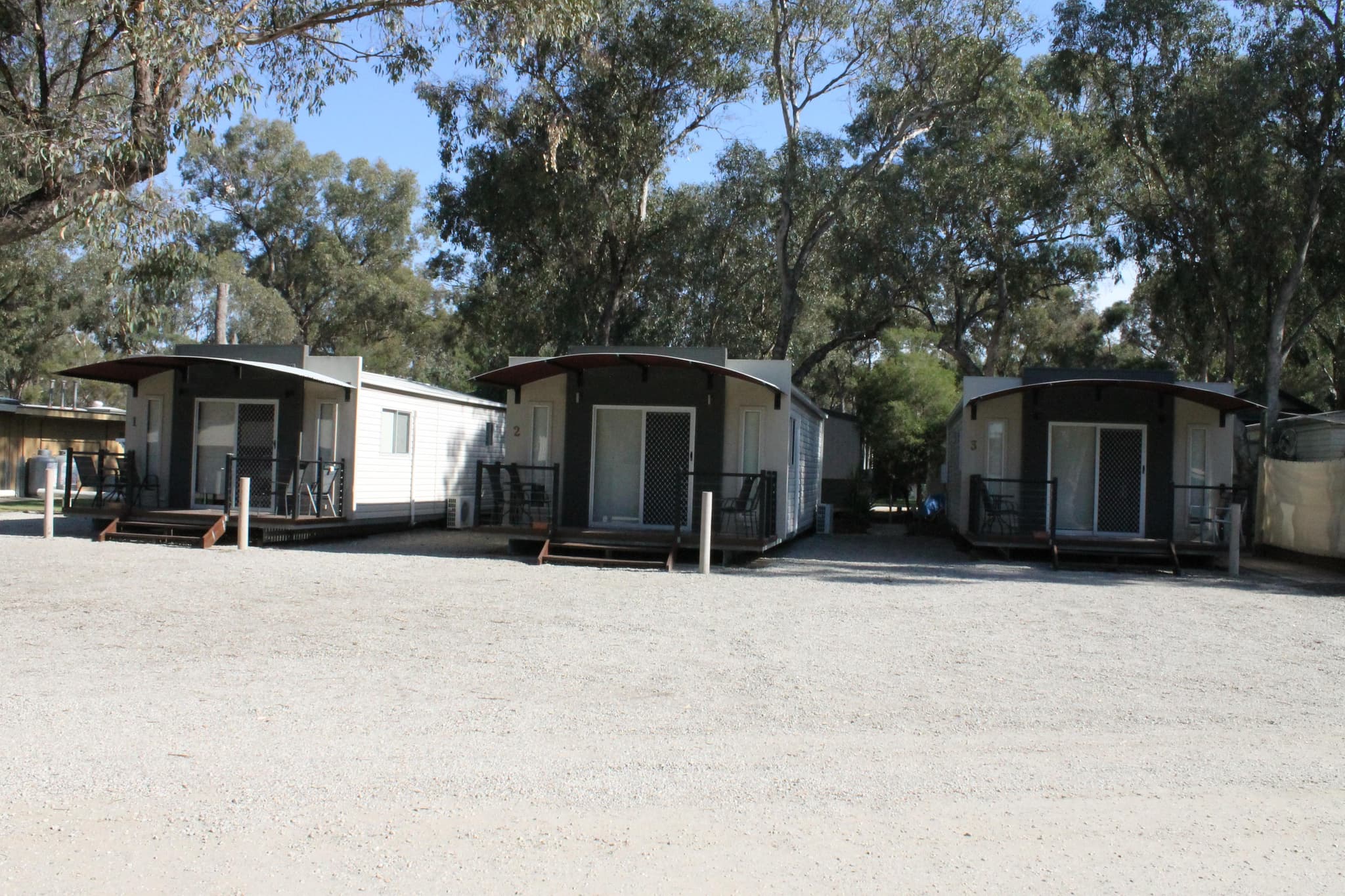 Numbered cabins with curved awnings and timber decks at Torrumbarry Weir Holiday Park, set among gum trees near Patho VIC