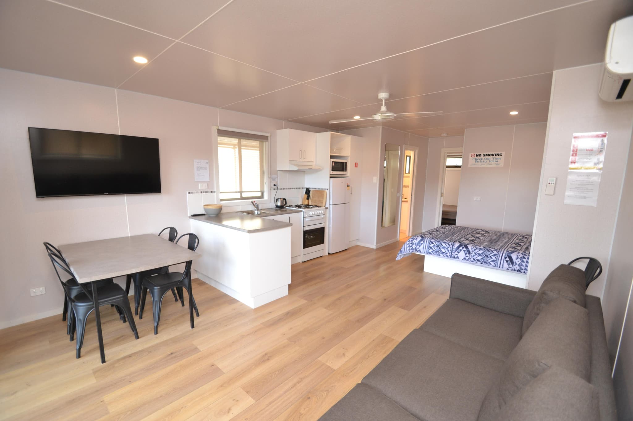 Open-plan cabin interior at Torrumbarry Weir Holiday Park with kitchen, dining table, wall-mounted TV, bed and grey lounge on timber flooring