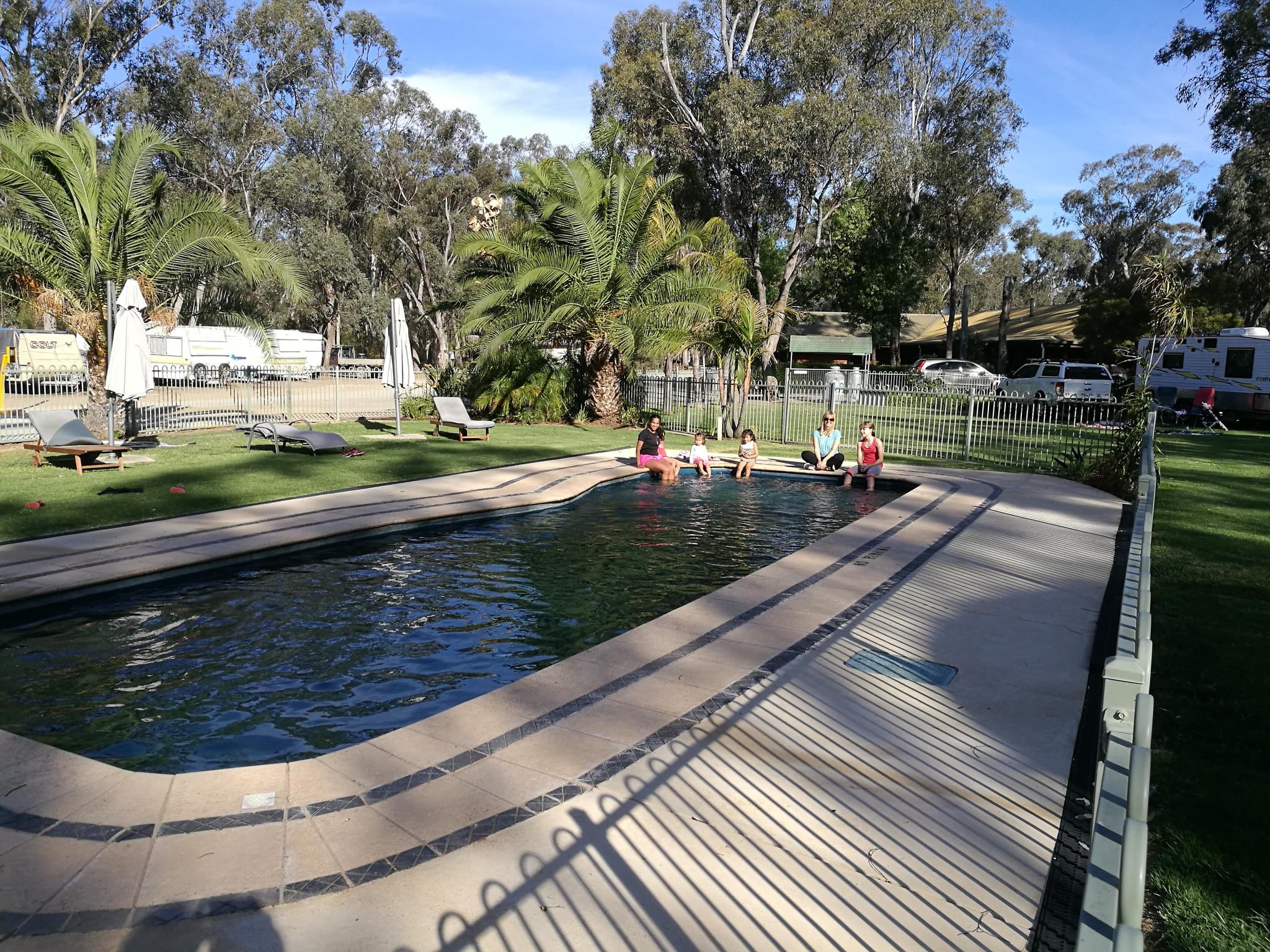 Fenced swimming pool at Torrumbarry Weir Holiday Park in Patho VIC, with families paddling and caravans parked among gum trees beyond the pool fence