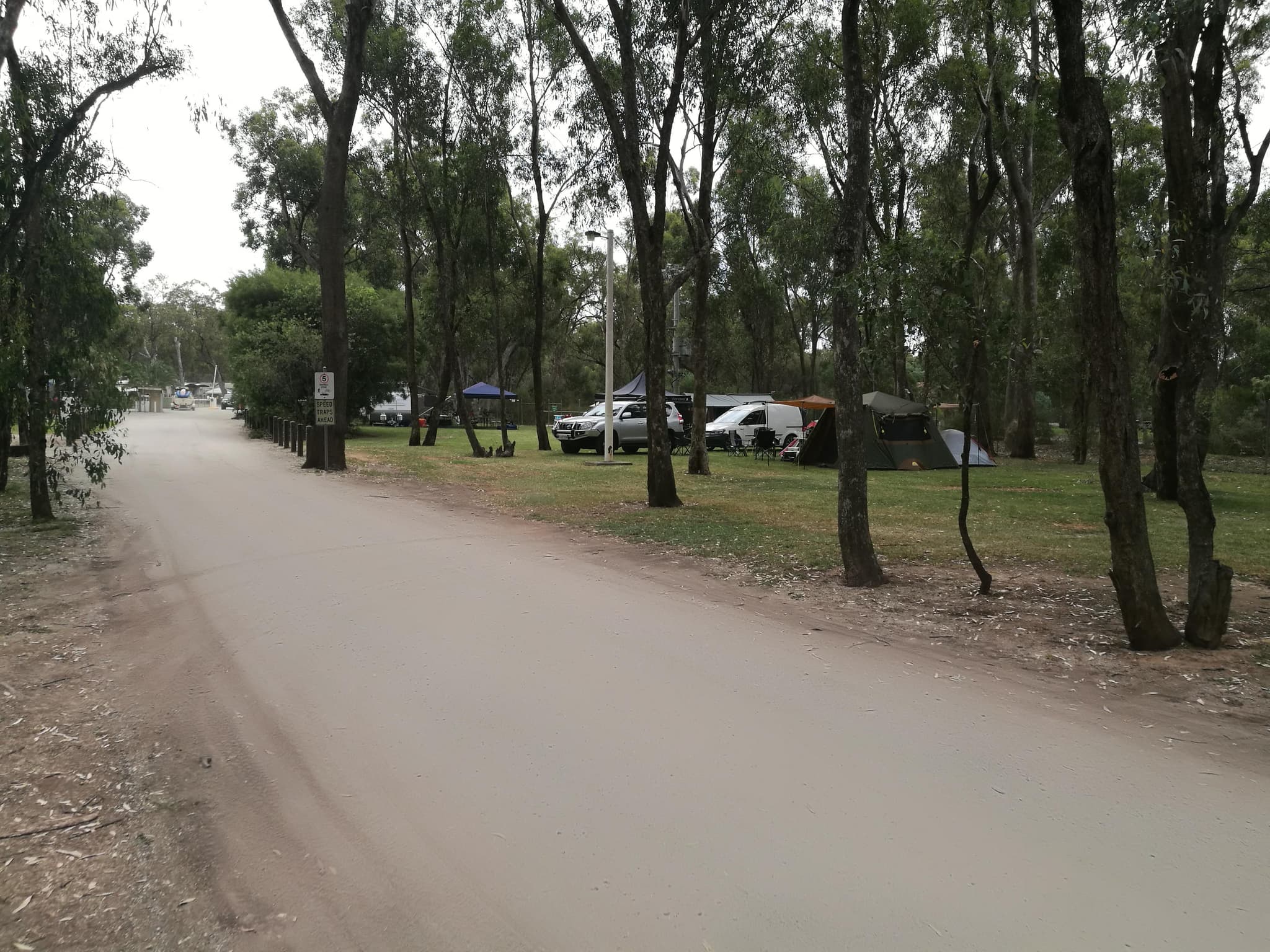 Grassy unpowered site under gum trees at Torrumbarry Weir Holiday Park, with tents, a 4WD and camping gear beside the dirt access road