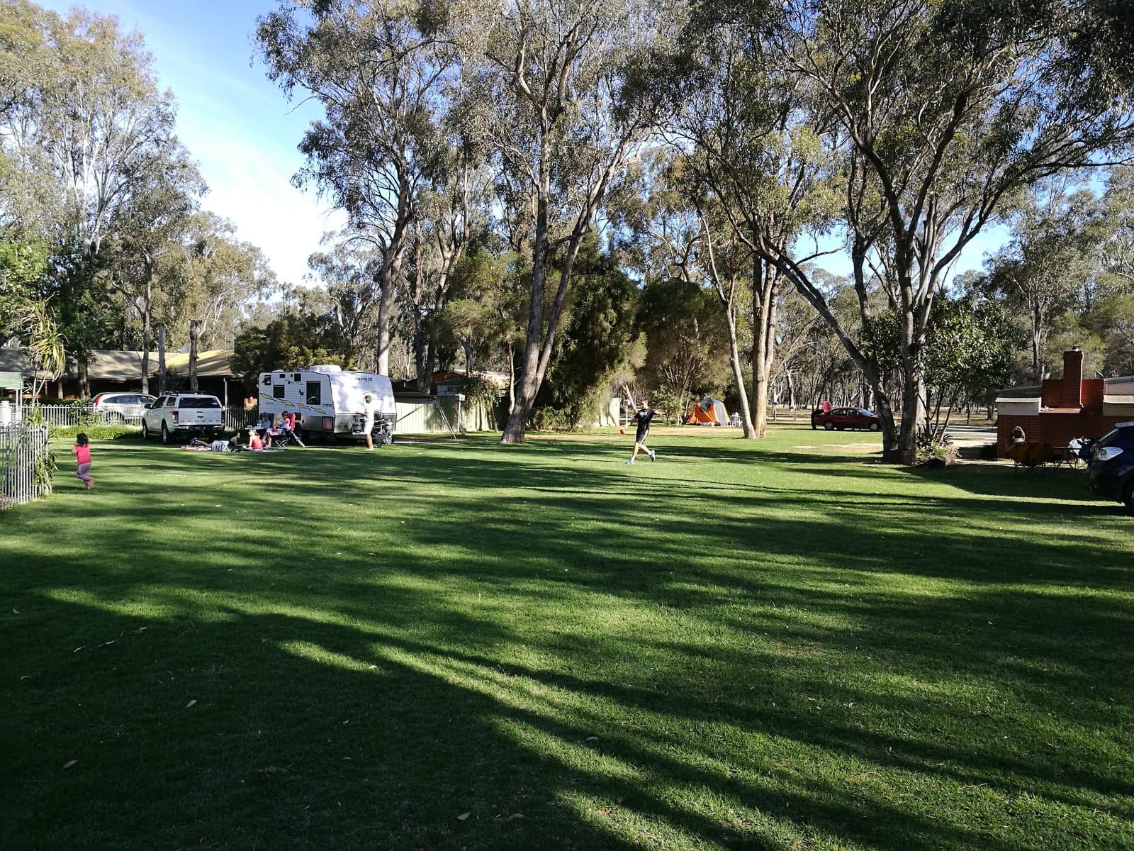 Grassy powered sites at Torrumbarry Weir Holiday Park, Patho VIC, with a caravan parked under tall gum trees and families relaxing on the lawn